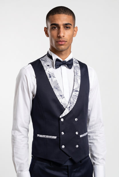 A man wearing a navy blue double-breasted waistcoat with a white and navy patterned, a white dress shirt, and a navy bow tie, photographed against a light gray studio background.